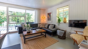 A modern living room with a sofa, coffee table, and TV. There are large windows and a view of greenery outside. - 4 Star Holiday Home in Norre Nebel (Norre Nebel)