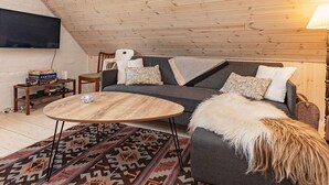 En hyggelig loftstue med træloft, en grå sofa med mønstrede puder, et rundt træbord og en vævet måtte.