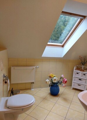 Une petite salle de bain avec un puits de lumière, des toilettes, un radiateur et un vase rempli de fleurs.