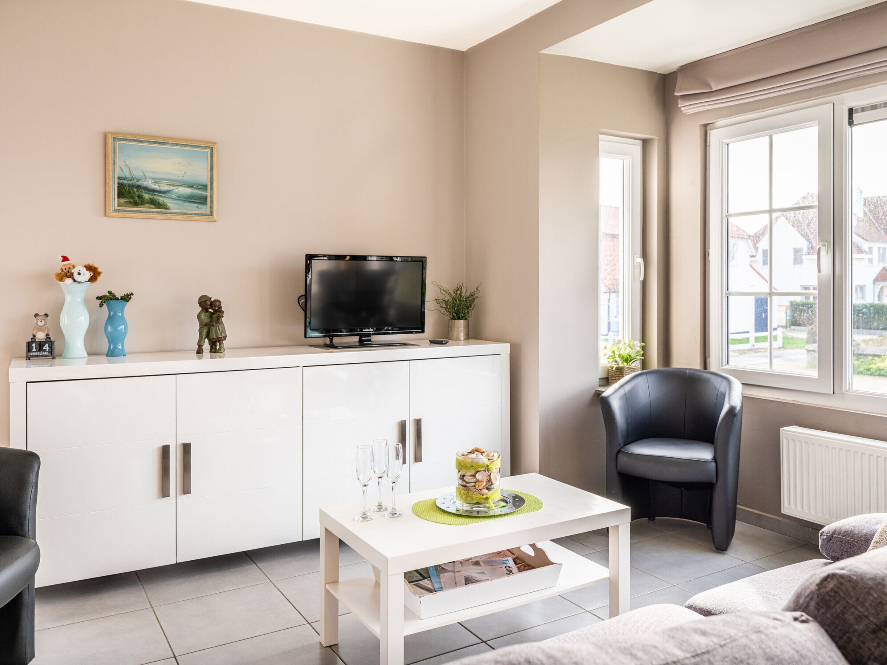 Un salon moderne avec un meuble blanc, un téléviseur à écran plat, un fauteuil en cuir noir et une fenêtre donnant sur des maisons.