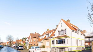 Une rue résidentielle bordée de maisons, avec des voitures garées et un ciel dégagé.