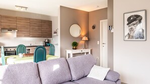 Un salon moderne avec un canapé gris, un coin repas avec une table et des chaises, une cuisine en arrière-plan et une horloge fixée au mur.