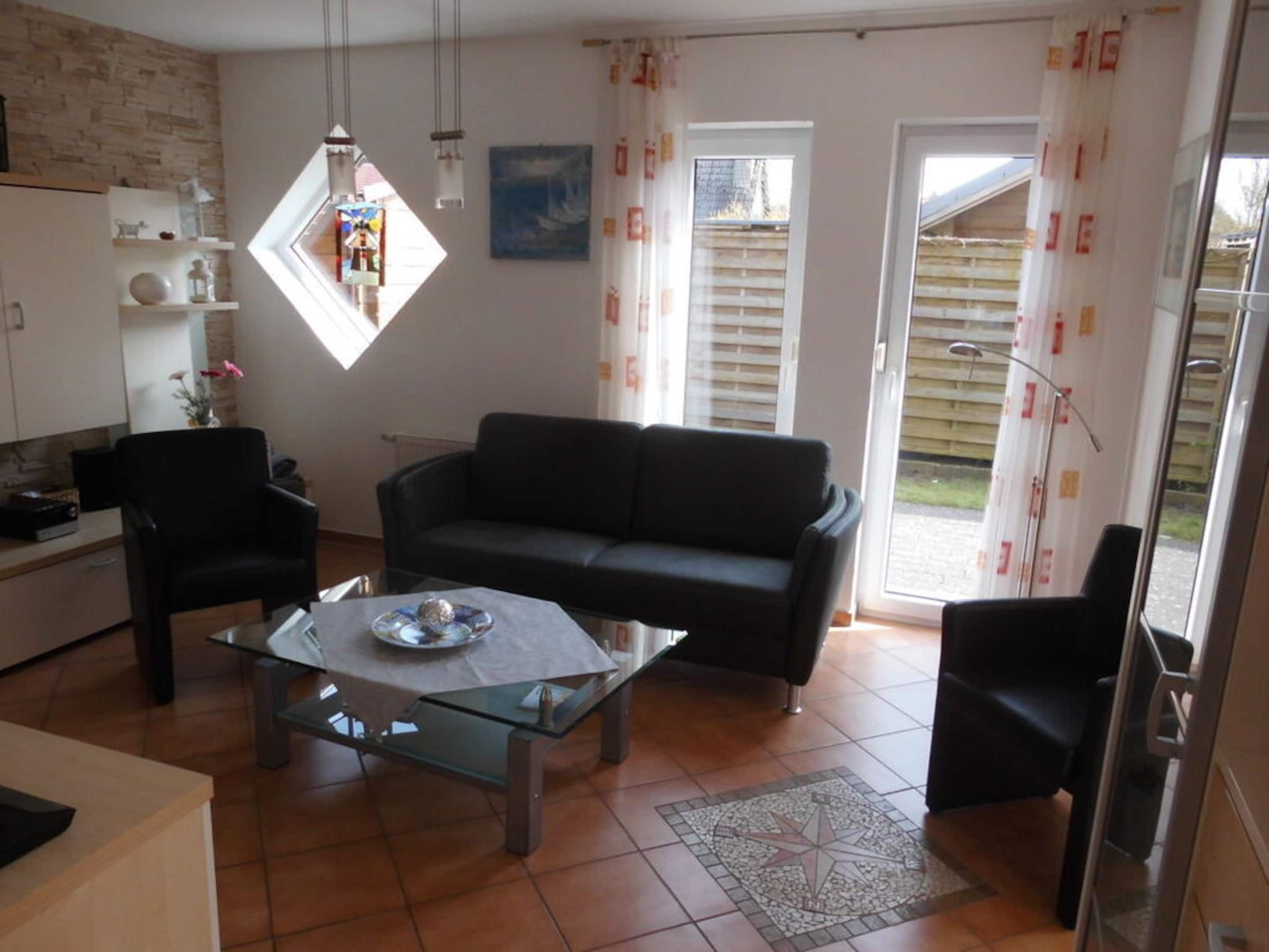 A living room with a black sofa, a glass coffee table, and a small dining table. There is a window with curtains, a radiator, and a wall-mounted shelf with decorative items.