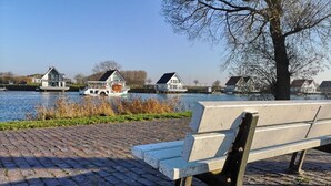 Een bankje langs een met bakstenen bestraat pad, met een rustig waterlichaam en huizen op de achtergrond.