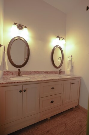A bathroom with two sinks, each with a round mirror and overhead lighting. There are cabinets below the sinks and a towel rack on the wall.