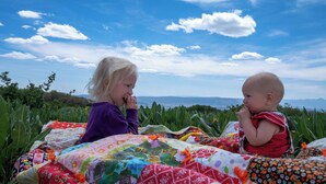 Dues nenes assegudes sobre una manta colorida a l'aire lliure, amb una vista panoràmica de muntanyes i vegetació al fons.