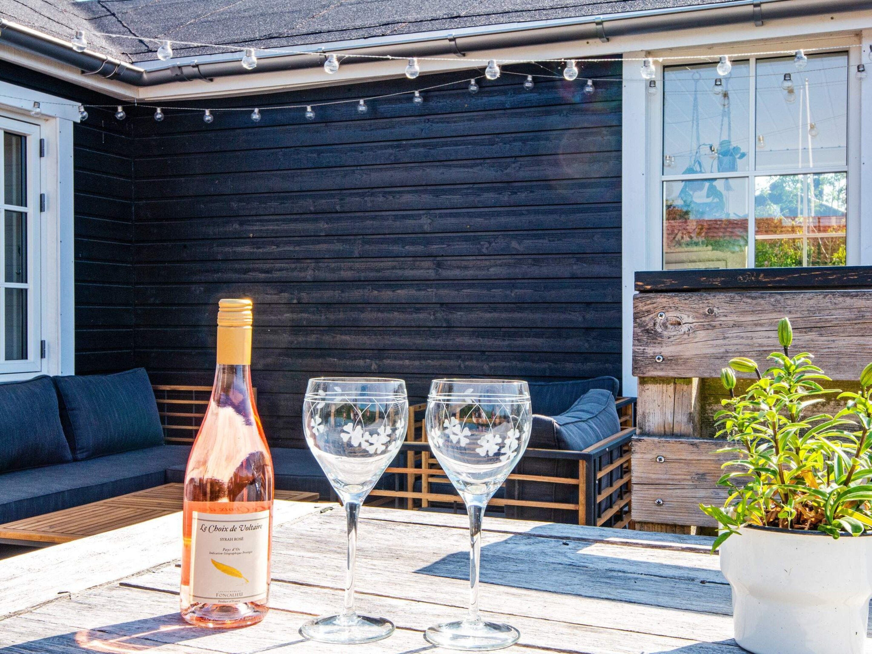 A bottle of wine, two wine glasses, a wooden bench, and a potted plant on an outdoor table.