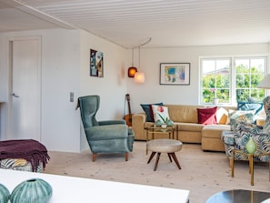 A living room with a sofa, armchair, ottoman, and a guitar. There are paintings on the walls and a window with a view. - 4 Person Holiday Home in Malling (Malling)