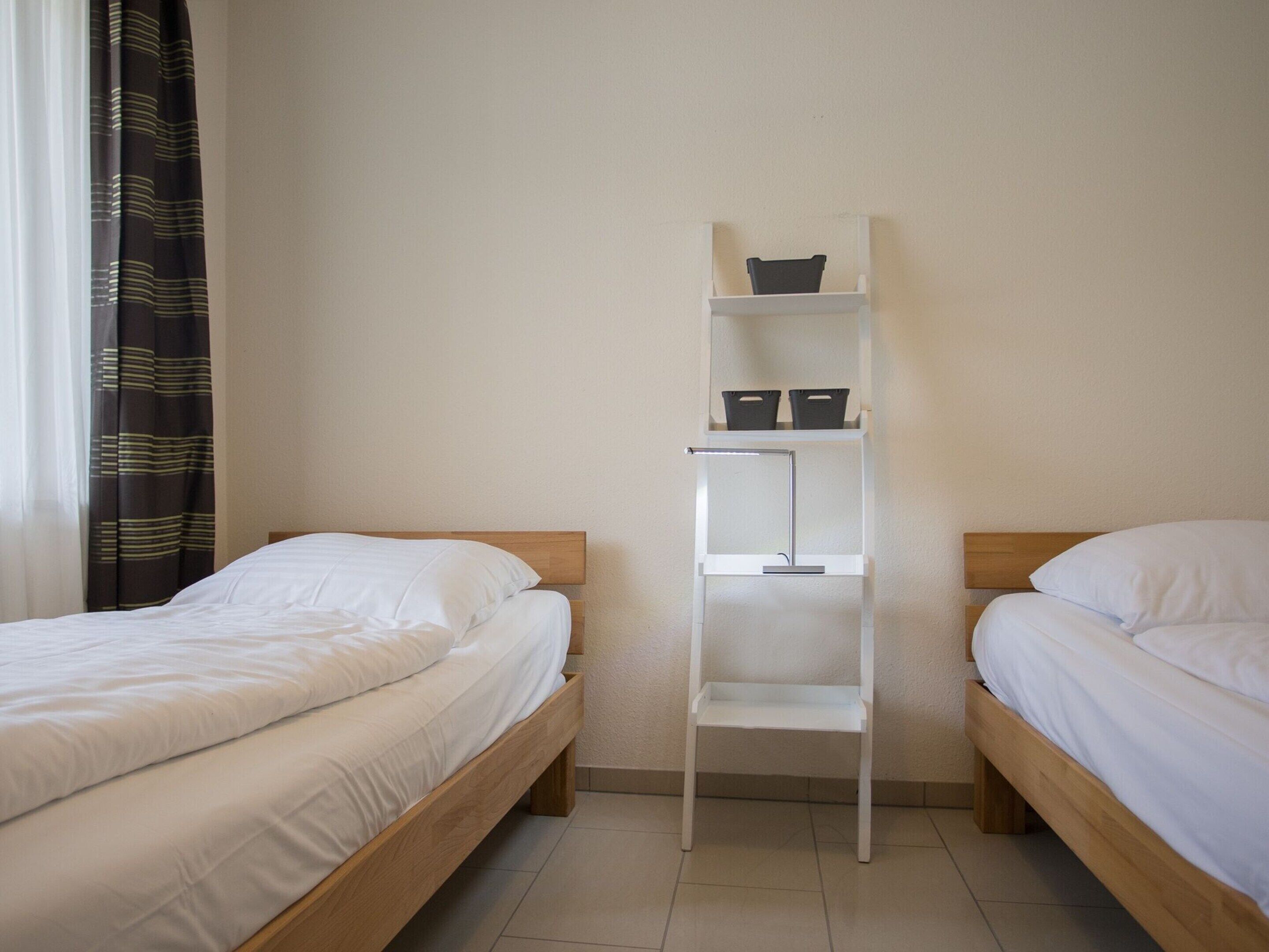 A room with two wooden beds, a white shelf unit, and a window with curtains.