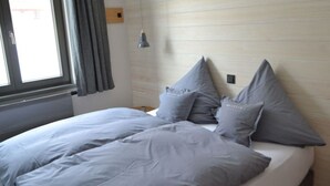 A neatly made bed with grey bedding and pillows, a window with curtains, and a wall-mounted light fixture. - 3 in het Huis Meerloft (Norderney)