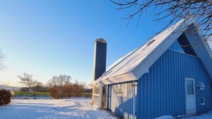 A blue building with a tall chimney, surrounded by snow-covered ground and sparse trees. - Holiday Home sea Waves (Brodersby)