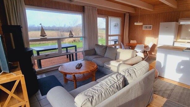 A cozy living room with a sectional sofa, a wooden coffee table, and a view of a garden through large windows. - Holiday Home sea Waves (Brodersby)