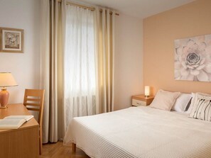 Une chambre d’hôtel comprenant un lit, un bureau avec une lampe, une chaise, une table de chevet avec une lampe et un mur orné d’un tableau représentant des motifs floraux.