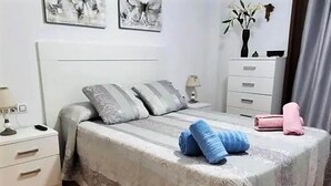 Een slaapkamer met een bed, kussens, een nachtkastje, een dressoir en een vaas met bloemen.