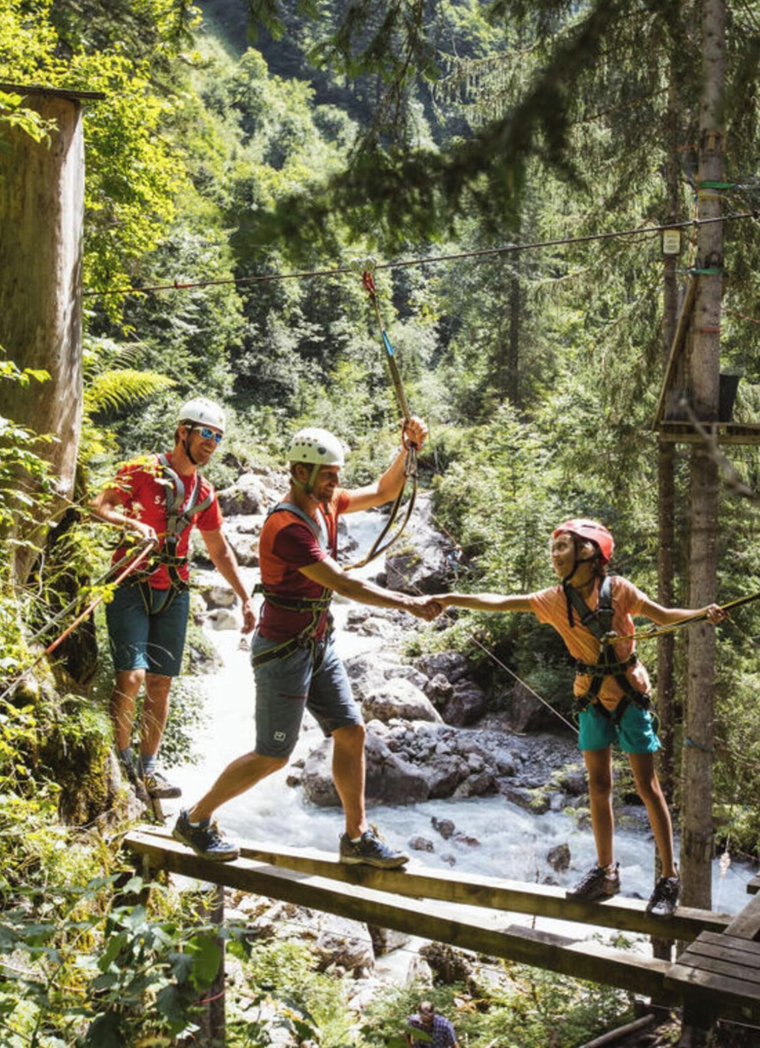 Trois personnes se trouvent sur un pont en bois, dans une zone boisée, au-dessus d’une rivière.