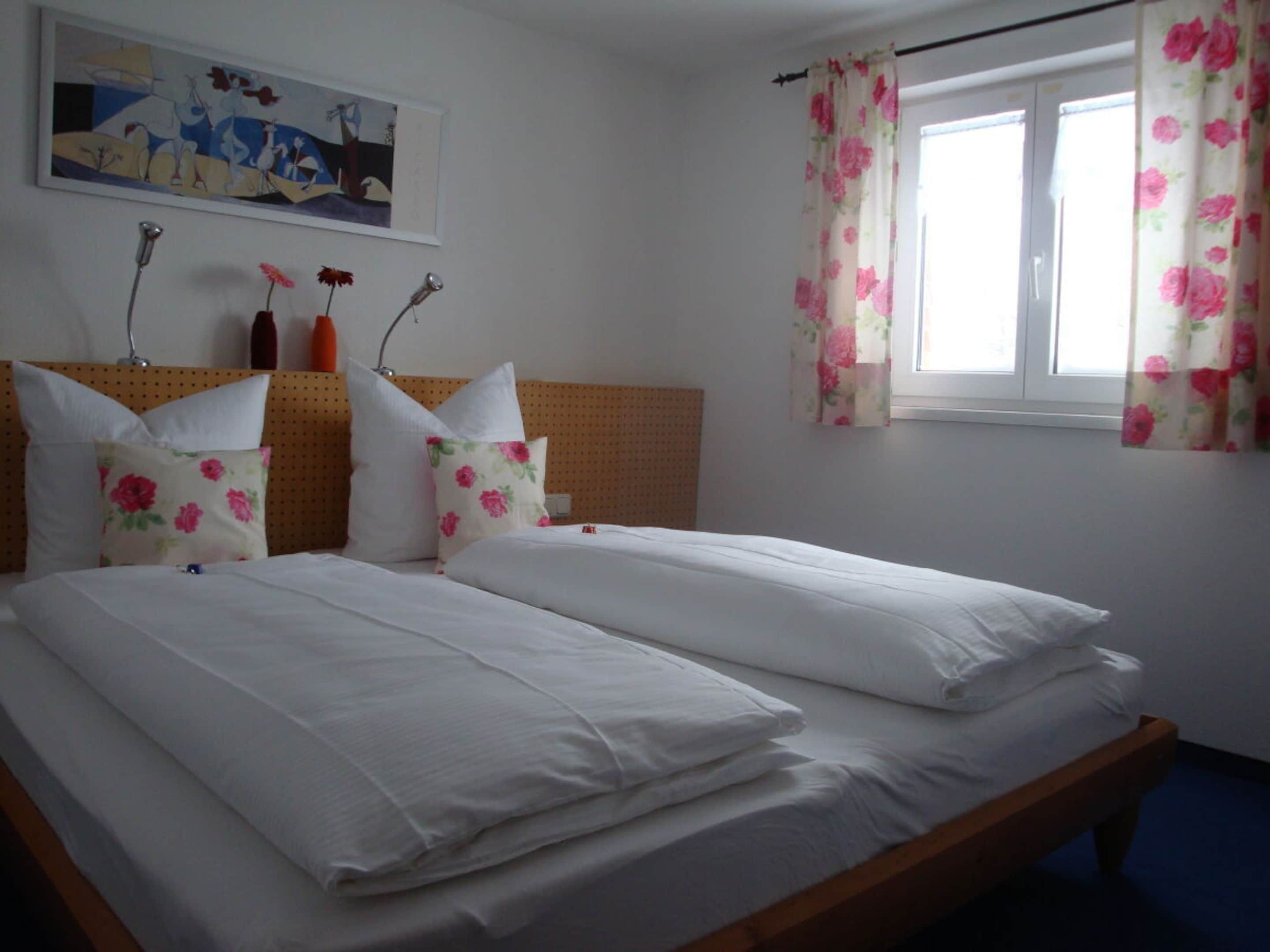 A double bed with white linens and floral-patterned pillows, a wooden headboard, a painting above the bed, and a window with floral curtains.