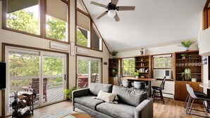 A spacious living room with a gray sofa, wooden coffee table, and a ceiling fan. Large windows offer views of the outdoors.