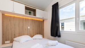 A neatly made bed with white linens and rolled towels, a wooden headboard, a small shelf with a plant, and a window with curtains. - Chalet in Middelkerke Near the Beach (Middelkerke)