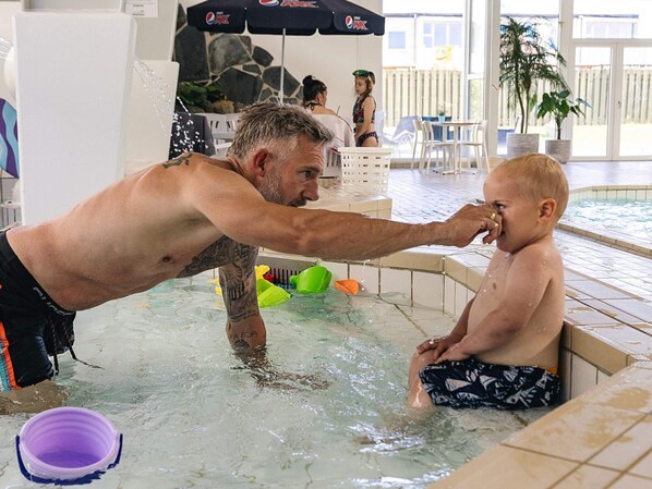 A man and a child in a pool with a stone wall and umbrellas in the background.