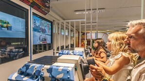 Et værelse med et stort skærm, der viser et computerspil, siddepladser med kasser i camouflage-mønster, og en person, der holder en spillekonsol.