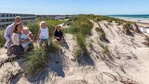 En familie på en sandstrand med klitter og en bygning i baggrunden.