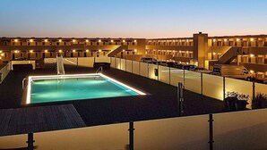 Un área de piscina en la azotea con una piscina rectangular, rodeada por un edificio de varios pisos con balcones y un paseo techado.