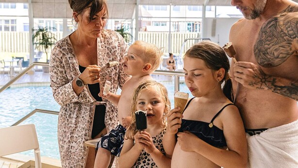 En familie ved en pool med krystalklart vand og et overdækket område med stole.