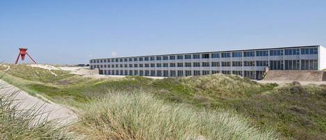 A modern building with a flat roof and large windows, surrounded by sand dunes and grass.