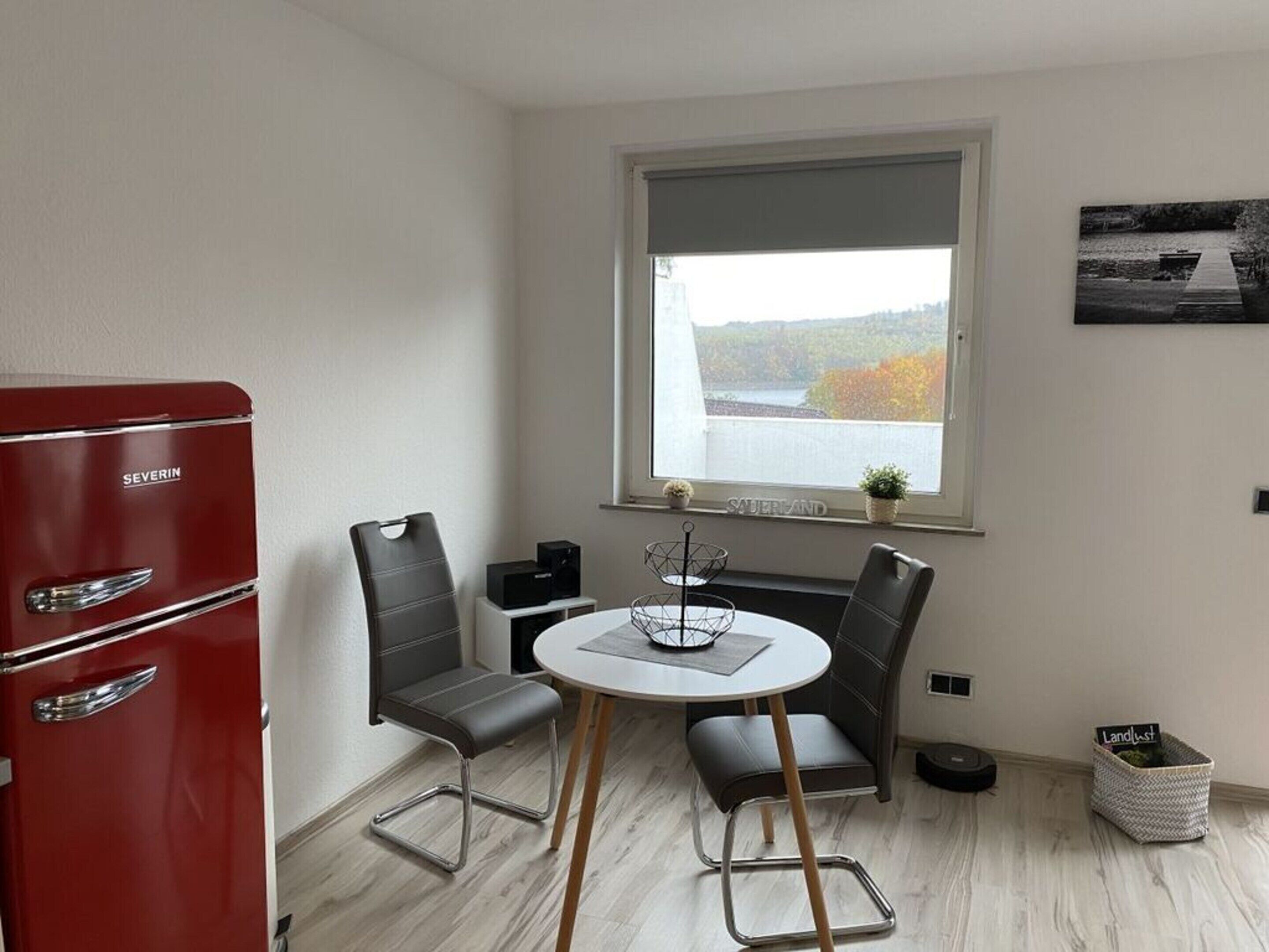A compact room with a red refrigerator, two chairs, a small table, and a window with a view of a lake.