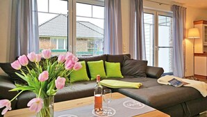 A modern living room with a sofa, a table with a vase of flowers, and a window with curtains. - Holiday Home sea Breeze (Butjadingen)