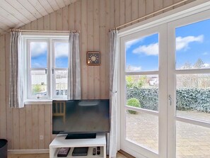 A room with a television on a white stand, a window with curtains, and a view of a garden. - 4 Person Holiday Home in Hemmet (Hemmet)