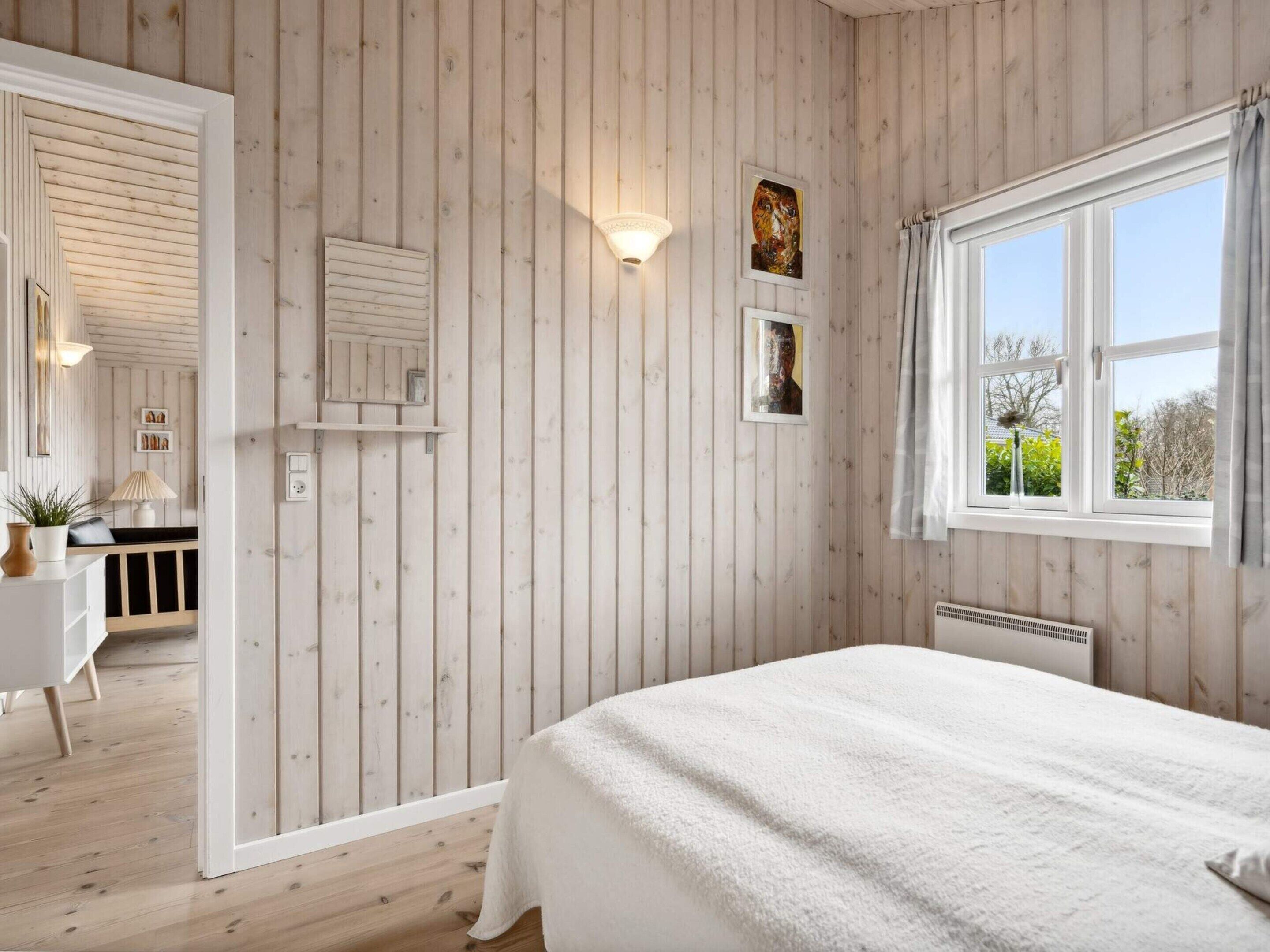 A bedroom with a large bed, wooden panel walls, a window with curtains, and a view of greenery outside.