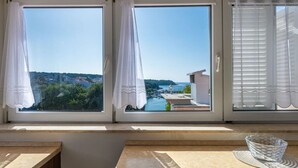 A room with three windows, each with white curtains. The windows offer a view of a coastal area with buildings and greenery. - Donatella -apt 2- met Zeezicht (Medulin)