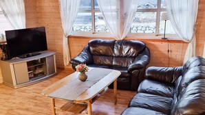 Un soggiorno con pavimento in legno, un divano in pelle nera, un tavolino in legno con un vaso di fiori, una TV su un mobile e una finestra con tende.
