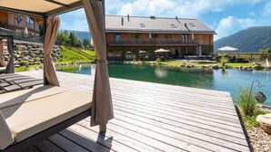 A wooden deck with a pool, a wooden building with balconies, and a mountain backdrop. - Chalet in Wolting With Natural Swimming Pond (Tamsweg)