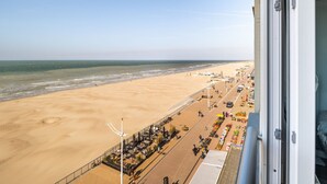 Une vue sur la plage depuis un balcon, avec une plage de sable, une promenade bordée de boutiques et un ciel dégagé.