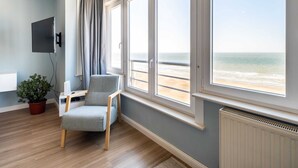 Une pièce dotée d’une grande fenêtre donnant sur une plage, d’un fauteuil gris, d’une petite table, d’une plante en pot et d’un téléviseur fixé au mur.