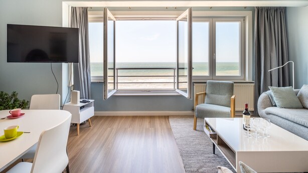 Un salon moderne doté d’une grande fenêtre donnant sur la mer, d’un téléviseur à écran plat, d’un coin repas et d’un espace détente avec un canapé et des fauteuils.