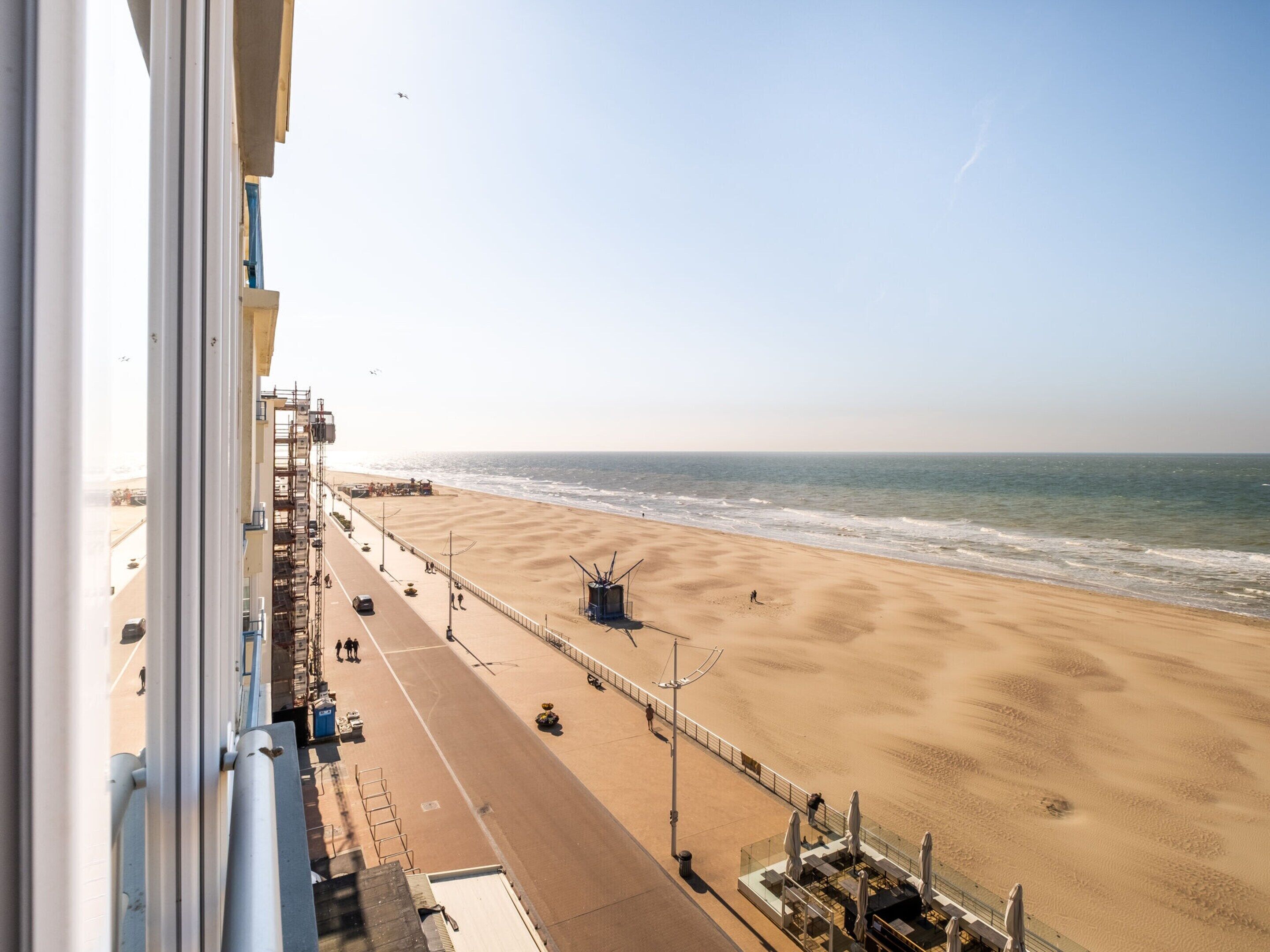 A beach view from a balcony with a clear sky, a sandy beach, and a road parallel to the beach.