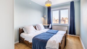 Une chambre à coucher avec deux lits en bois, un bureau, une chaise, une fenêtre avec des rideaux et un luminaire suspendu.