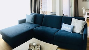 A modern living room with a dark blue sectional sofa, two light blue pillows, a small table with a candle and a decorative item, and a view of a dining area through a glass door.