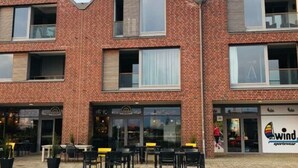 A brick building with a flat roof and large windows. There are outdoor tables and chairs in front.
