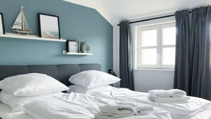 A neatly made bed with white linens and towels, a headboard, a window with curtains, and a shelf with decorative items. - Comfortplus Apartment With 1 Bedroom (Dranske)