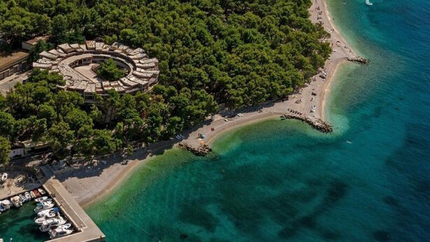 An aerial view of a coastal area with a circular building surrounded by dense trees, a sandy beach, and clear blue waters. - Apartments Pezo - Superior Two Bedroom Apartment With Balcony (Baska Voda)