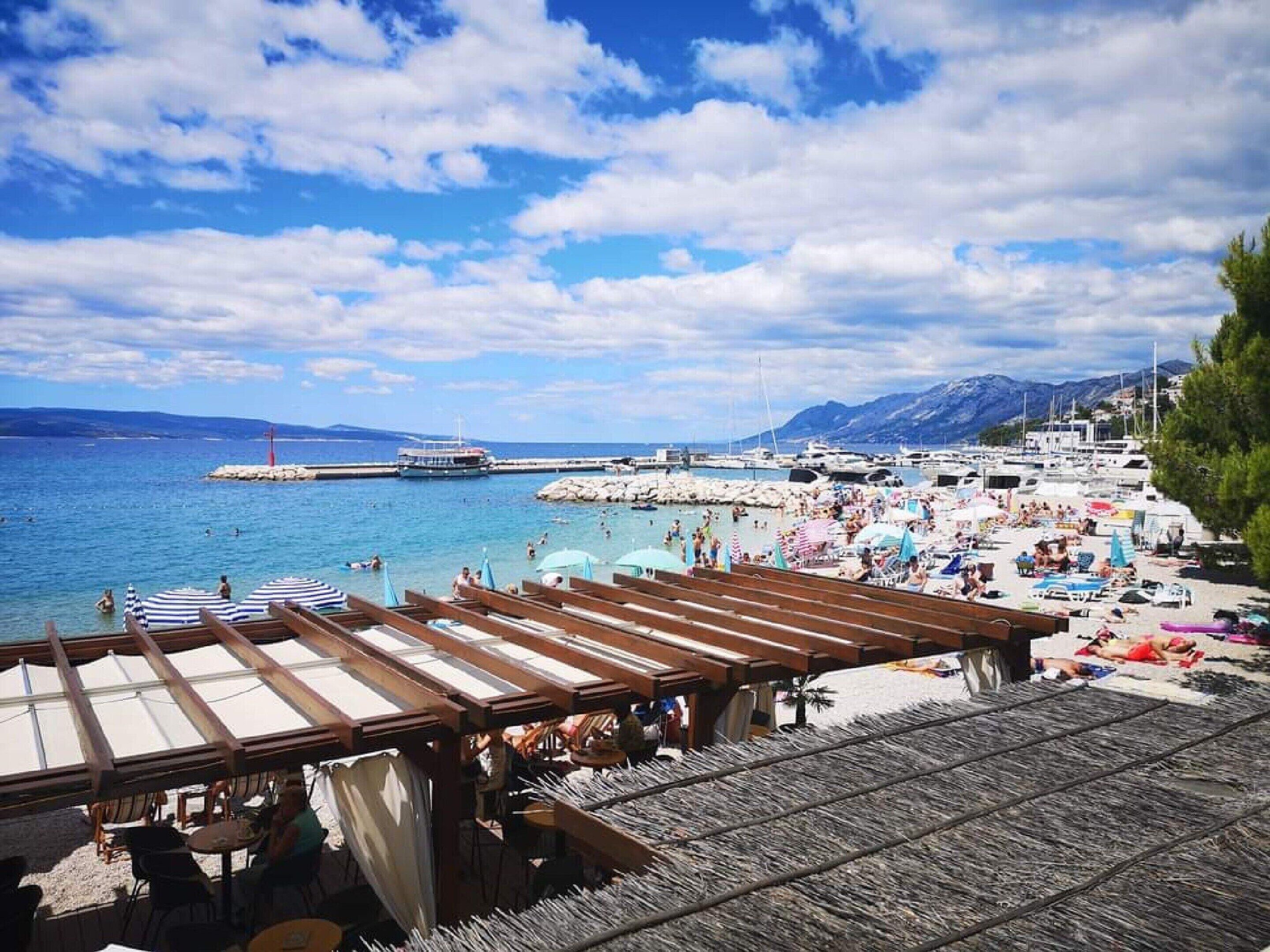 A beachside view with wooden deck seating, umbrellas, and a marina in the background.