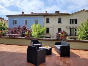 A rooftop terrace with outdoor furniture, a view of a building with multiple windows, and potted plants.