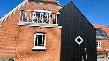 Un edificio de ladrillo rojo con un añadido de madera negra, un balcón con barandillas blancas y una ventana pequeña.