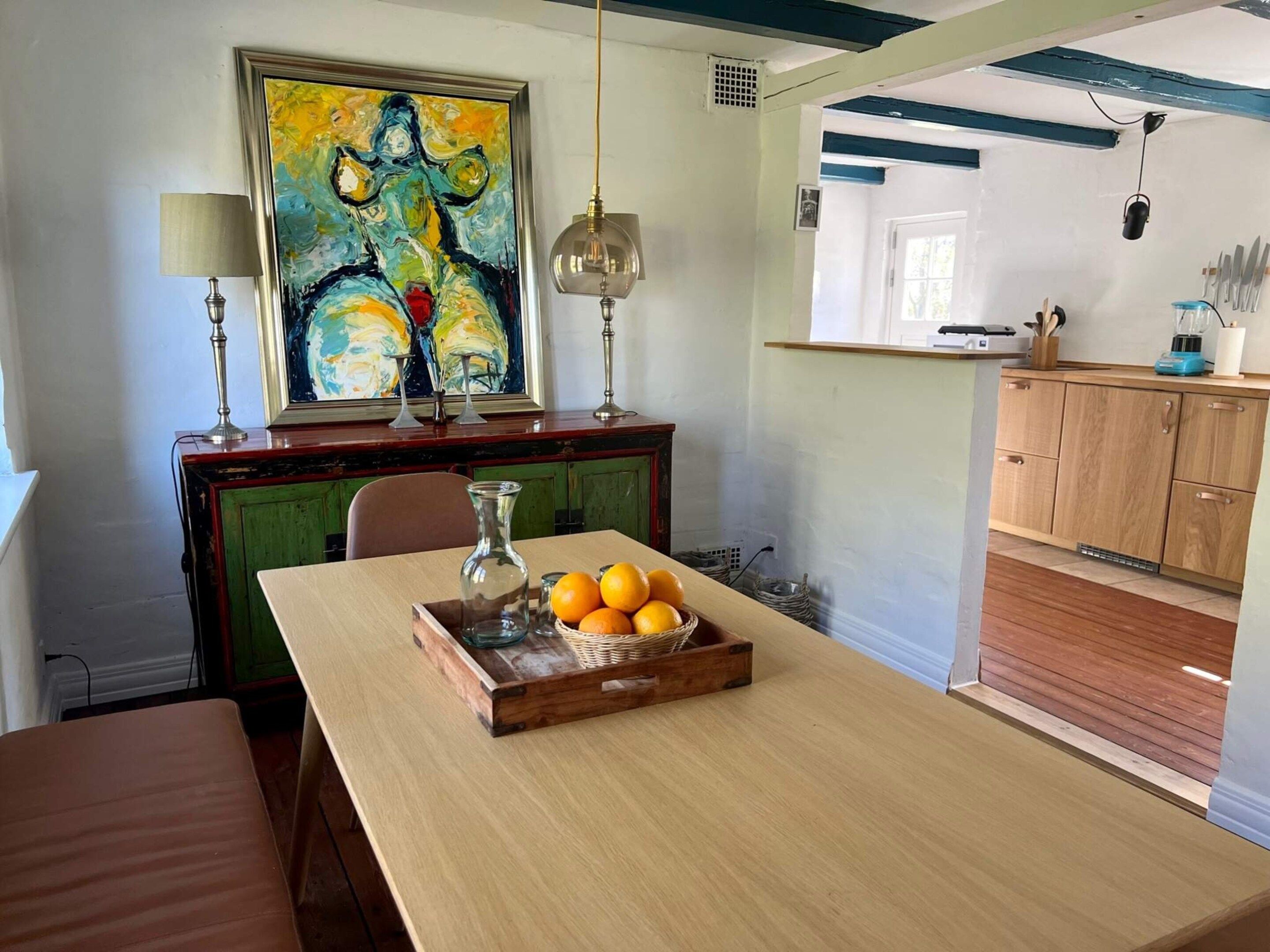 A dining area with a table set for two, a painting above the table, and a kitchen area visible through an open door.