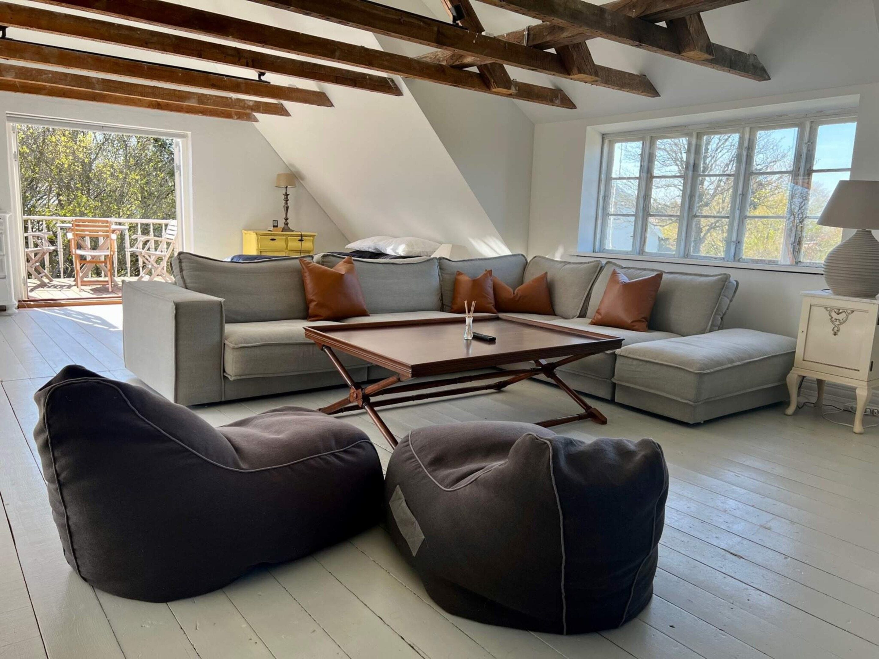 A modern loft-style living room with a high ceiling, exposed wooden beams, a sectional sofa, two bean bags, a coffee table, and a view of the outdoors.