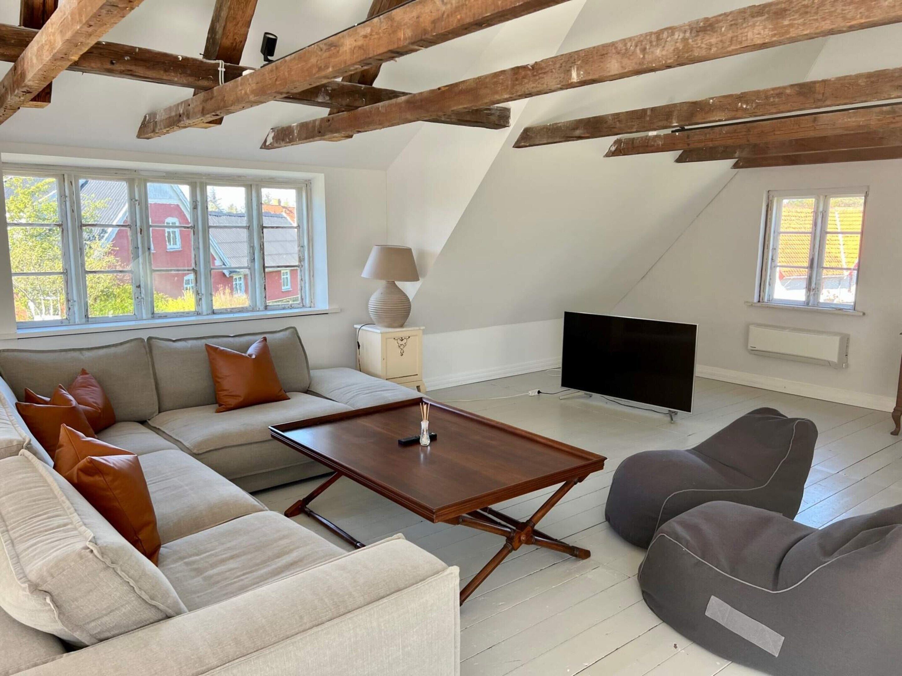 A loft-style living room with exposed wooden beams, a sofa set, a coffee table, a TV, and large windows with a view of buildings outside.